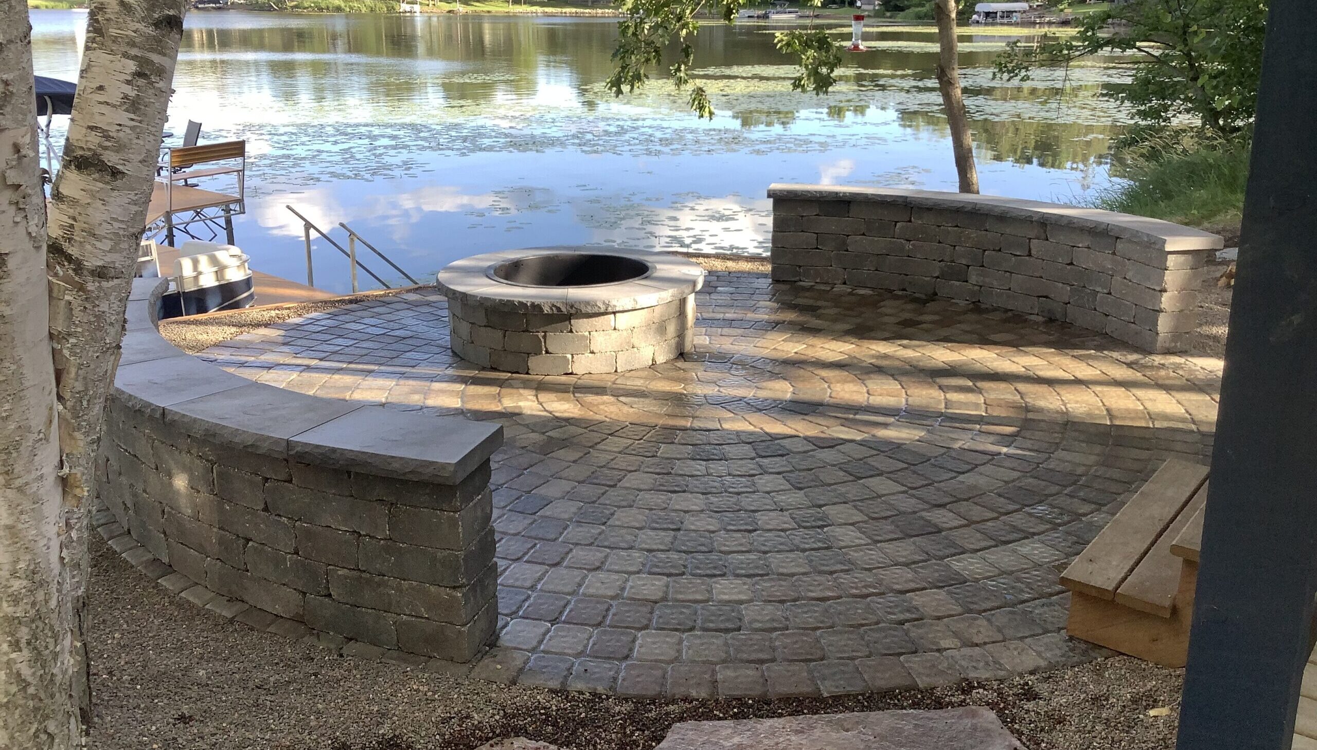 A large fire pit with stone pavers around it and bench seating for guests to enjoy. Lakeside landscaping done right by the Landscape Guys in Minneapolis, MN. 